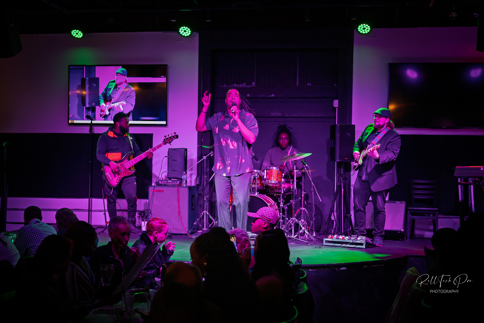 A live music performance with a singer and band on stage, including a bassist, drummer, and guitarist. The seated audience enjoys the show, creating a lively atmosphere.