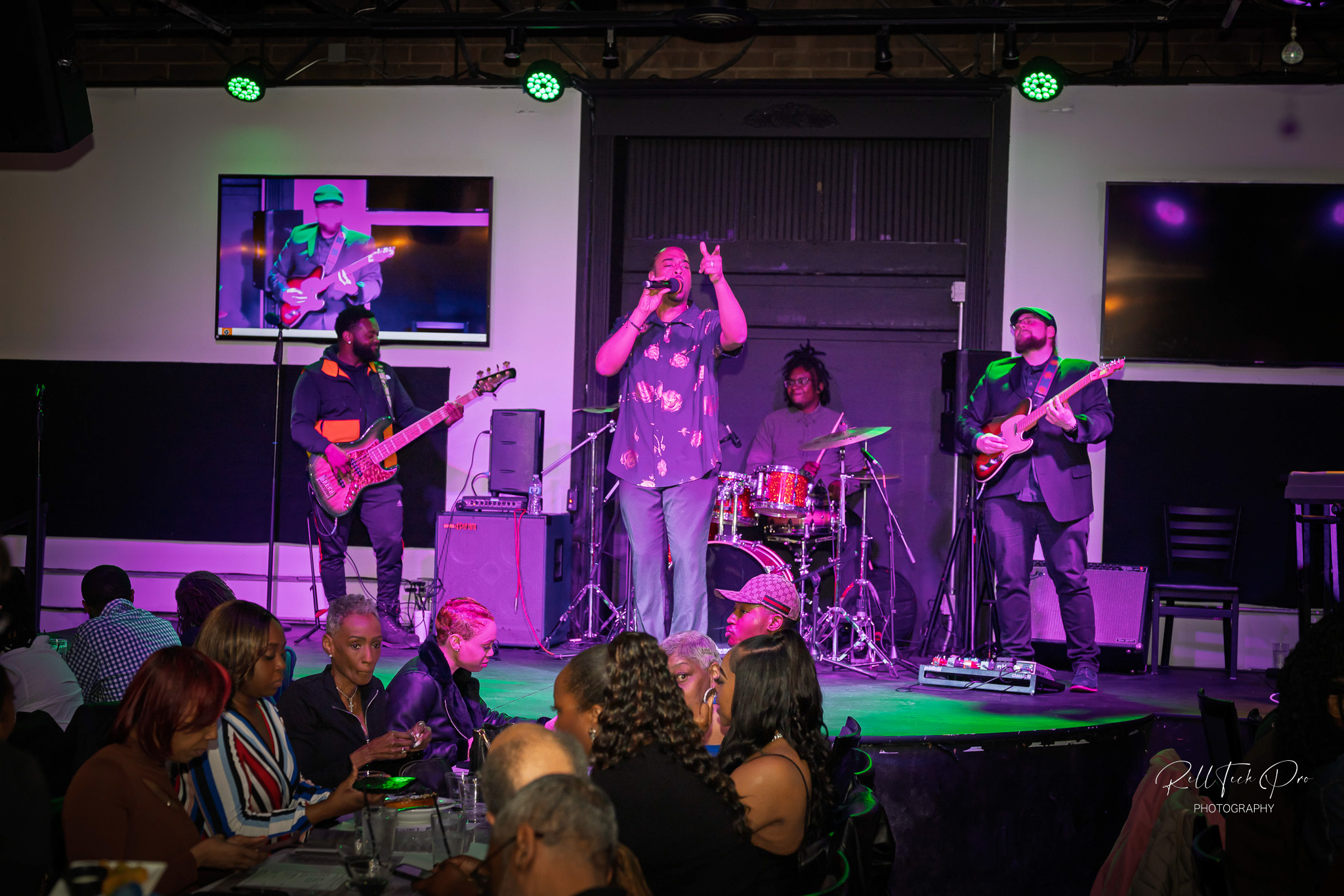 A live music performance with a singer and band on stage, including a bassist, drummer, and guitarist. The seated audience enjoys the show, creating a lively atmosphere.