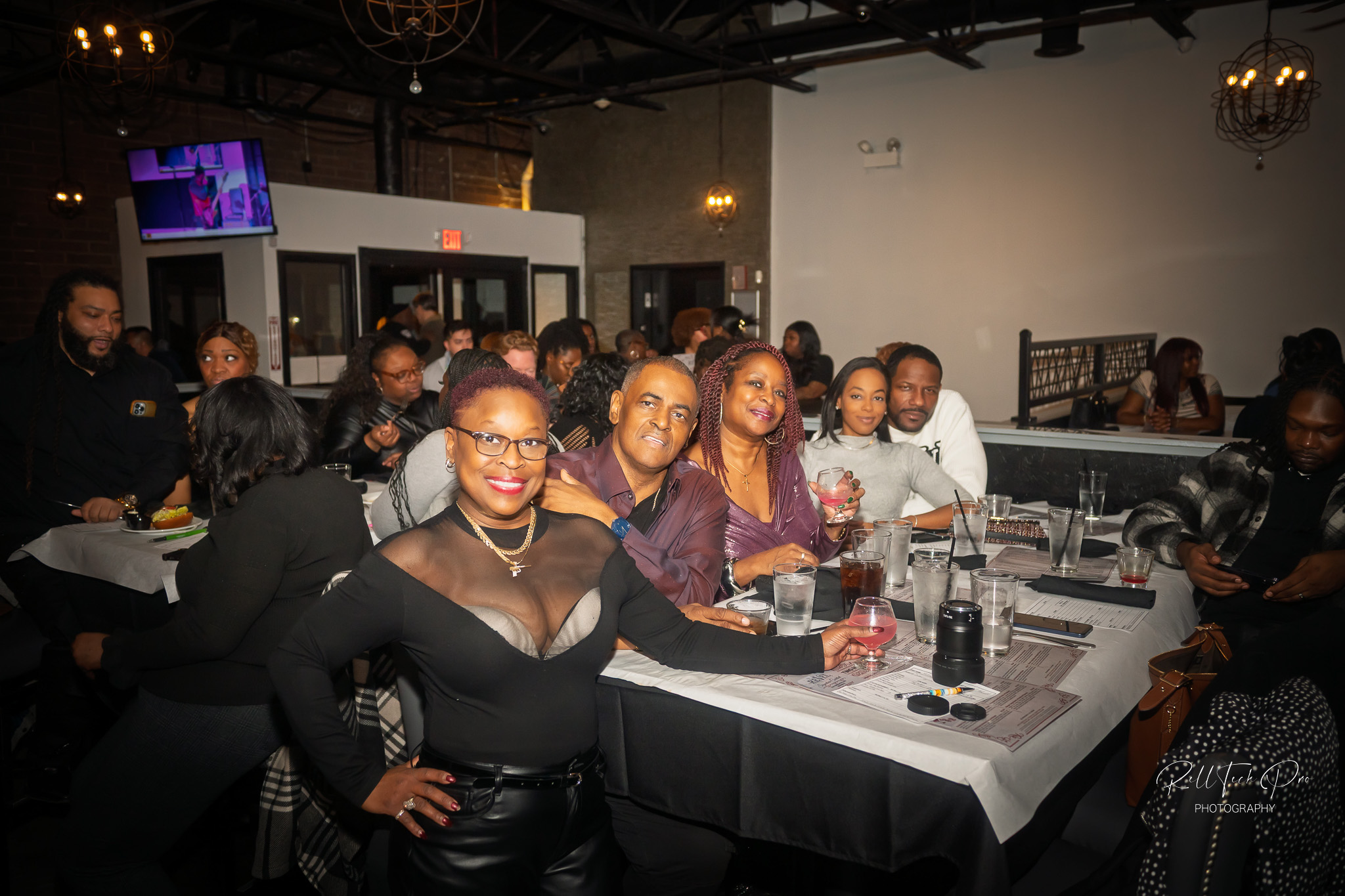 a group of people enjoying a meal together at a lively social event. One woman, smiling confidently, is posing in the front with a glass in hand. The group is seated at a large table with drinks and food, and the background reveals more guests enjoying the atmosphere. The setting appears to be in a stylish and relaxed restaurant or bar.