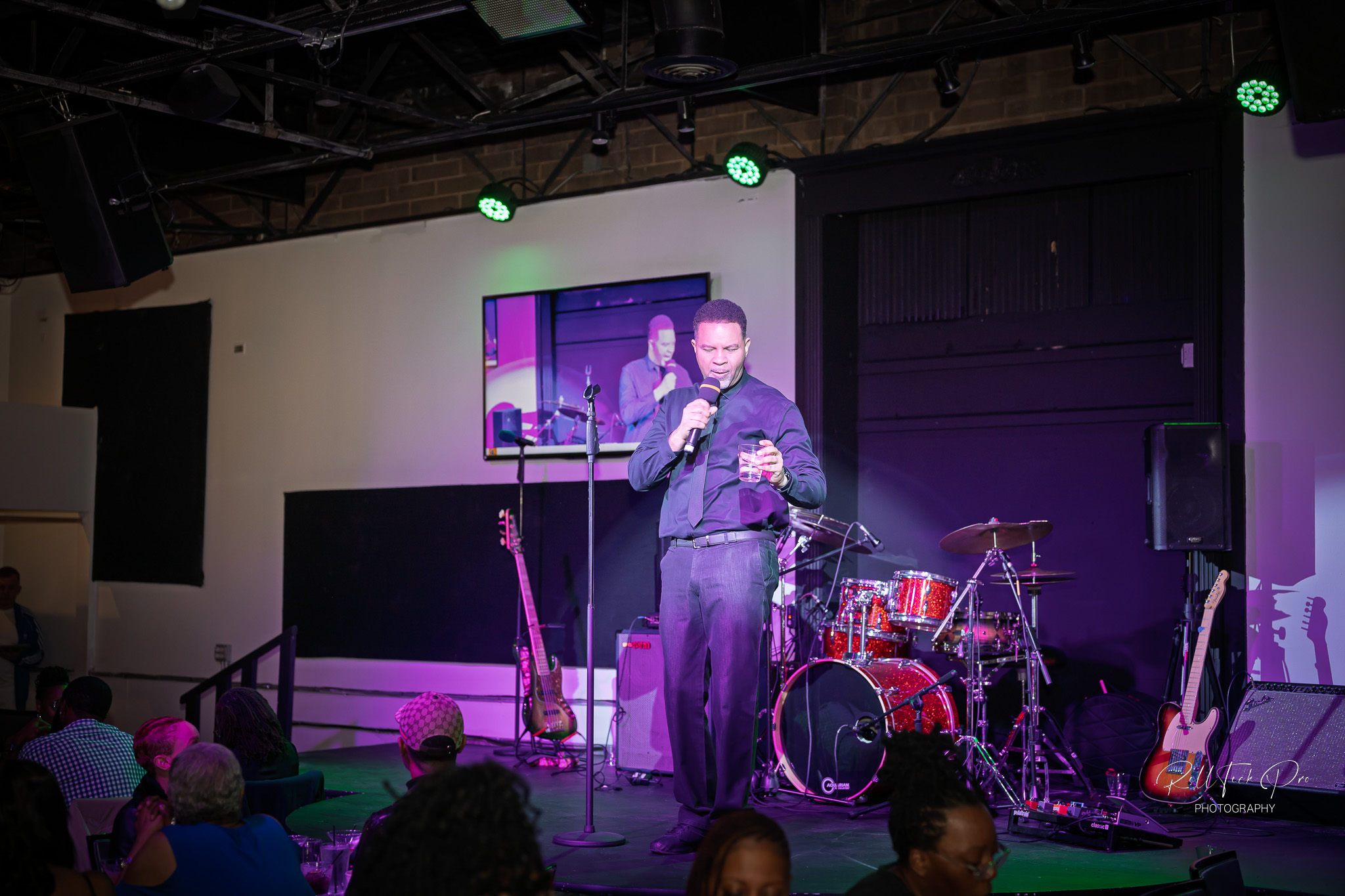 Vibrant live music featuring drums, guitar, and keyboard. The engaged crowd enjoys the performance, with some seated at tables, dining and socializing, all enhanced by colorful lighting.