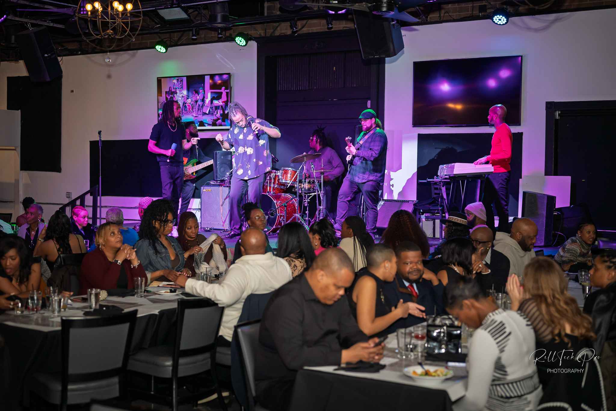 Vibrant live music featuring drums, guitar, and keyboard. The engaged crowd enjoys the performance, with some seated at tables, dining and socializing, all enhanced by colorful lighting.