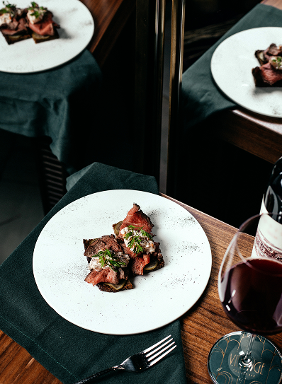 a beautifully presented plate of roasted meat with garnishes, possibly served with a side of vegetables or sauce, on a minimalist white plate. A glass of red wine is placed beside it, and the elegant setting is complemented by the rich dark tones in the background, which include a reflection in a nearby mirror. The table is neatly set with a green napkin and silverware, creating a refined atmosphere.
